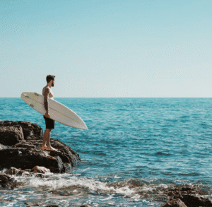 Young man with surfboard standing on rocks by the ocean.